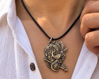 Puede incluir: Un colgante de tono bronce que presenta un relieve detallado de un rostro adornado con un motivo de pluma de pavo real y una flauta, suspendido de un sencillo collar de cordón negro. El colgante muestra texturas intrincadas y un acabado metálico.