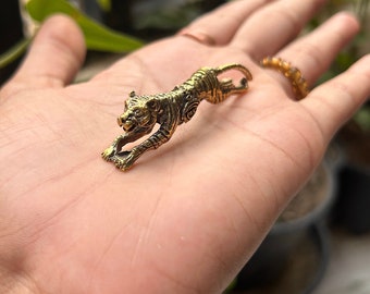 May include: A small, detailed brass tiger figurine in a running pose. The tiger is a golden color with intricate etched details, including stripes and facial features. The figurine is held in a hand, with a blurred background of greenery.
