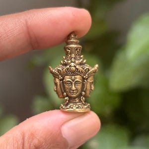 Puede incluir: Un colgante de metal dorado con tres caras de una deidad. El colgante está intrincadamente detallado y tiene una parte superior con forma de corona.