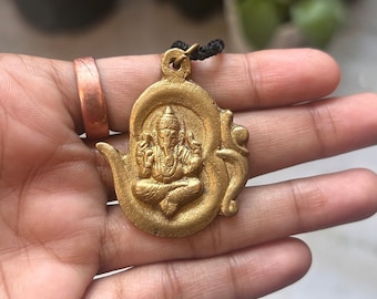 May include: Gold-toned pendant necklace featuring a detailed depiction of the Hindu deity Ganesha, sitting in a meditative pose within a stylised Om symbol.