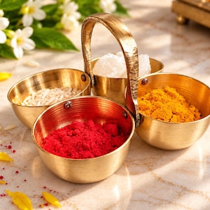 May include: A brass spice container featuring three small, rounded bowls connected by a central arched handle. The bowls contain white rice grains, white sugar crystals, bright yellow turmeric powder, and vibrant red powder.