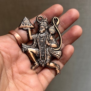 Könnte beinhalten: Ein bronzefarbener Metallanhänger, der die hinduistische Gottheit Hanuman darstellt, der in seiner linken Hand einen Berg und in seiner rechten Hand eine Keule hält. Der Anhänger befindet sich in einer Sprungpose mit einem Schwanz, der um seinen Körper geschlungen ist.