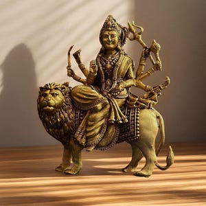 May include: A golden-colored statue of a deity riding a lion. The deity has multiple arms and is adorned with jewelry and a headdress. The lion is detailed with a textured mane and is in a walking pose. The statue is on a wooden surface.