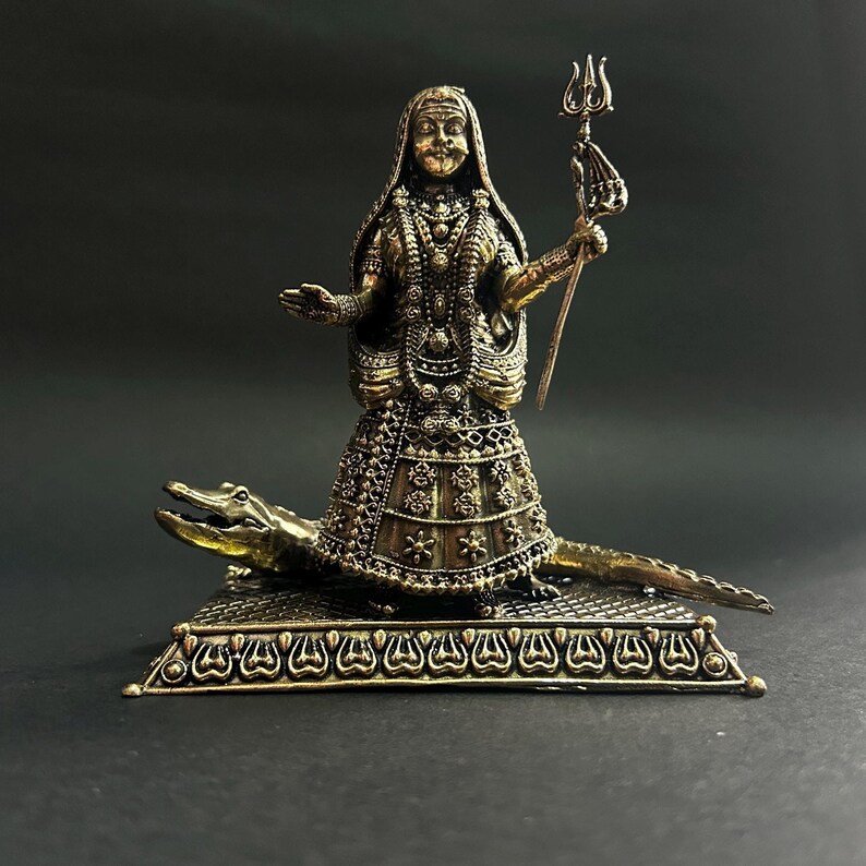 K&ouml;nnte beinhalten: Detaillierte Bronzestatue einer weiblichen Figur, geschm&uuml;ckt mit aufwendigem Schmuck und einem gemusterten Kleid, das einen Dreizack h&auml;lt. Die Figur steht auf einem Krokodil auf einem dekorativen Sockel. Die Statue hat eine dunkelbronze Oberfl&auml;che.