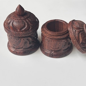 May include: Two intricately carved wooden boxes with lids. The boxes are dark brown and have a detailed design of flowers and leaves. The lids are slightly domed and have a pointed top.
