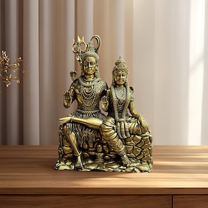 May include: A golden-colored statue depicting a seated couple, likely deities, adorned with intricate jewelry and headdresses. The figures are seated on a textured base, with one holding a trident. The statue is placed on a wooden surface.