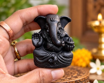 Op de afbeelding: Een klein, matzwart beeldje van de godheid Ganesha in zittende houding. Het figuur heeft grote oren, een gekrulde slurf en een opgeheven hand in een zegenend gebaar, met een kleine kom in de andere hand, alles uitgevoerd in donkere steen.