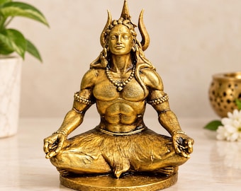 May include: A gold-colored statue of a deity in a seated meditative pose. The figure has long hair, a crescent moon on the head, and wears beaded necklaces and armbands. The statue is positioned on a circular base with a textured finish.