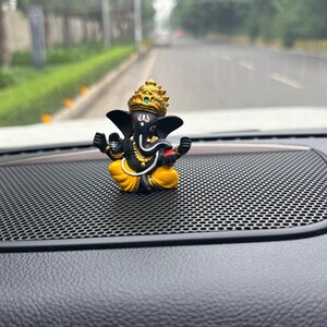 Puede incluir: Una peque&ntilde;a figura negra y dorada del dios hind&uacute; Ganesha, sentado en una postura meditativa con la trompa levantada. La figura est&aacute; colocada en un tablero de instrumentos de coche negro con una superficie de panal.