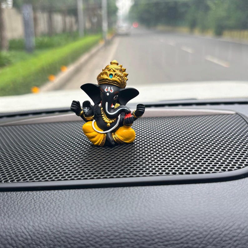 Puede incluir: Una peque&ntilde;a figura negra y dorada del dios hind&uacute; Ganesha, sentado en una postura meditativa con la trompa levantada. La figura est&aacute; colocada en un tablero de instrumentos de coche negro con una superficie de panal.