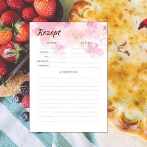 May include: A printable recipe card with a watercolour background featuring pink flowers. The card has sections for the date, ingredients, and preparation instructions. The card is surrounded by a table setting with strawberries, blueberries, and a pie.