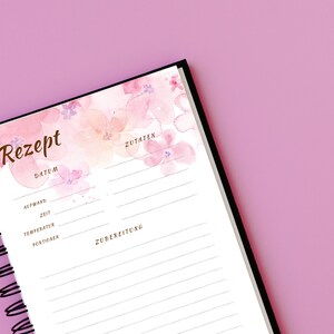 May include: A white recipe journal with a black spiral binding. The cover features a watercolour design of pink and peach flowers. The journal has sections for the recipe name, date, ingredients, and preparation instructions.