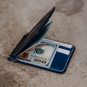 Può includere: Un portafoglio in pelle blu con cuciture bianche, contenente una banconota da cento dollari. Il portafoglio ha un fermasoldi e fessure per carte. Il design è minimalista e moderno.