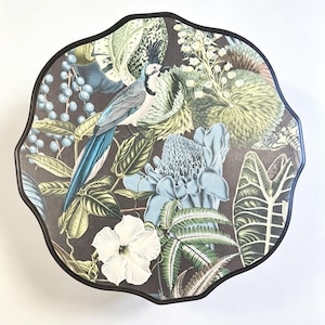 May include: A round table with a scalloped edge and a dark brown frame. The tabletop features a detailed botanical print on a dark background, showcasing a blue and grey bird, large light blue flowers, green ferns, and various tropical leaves.