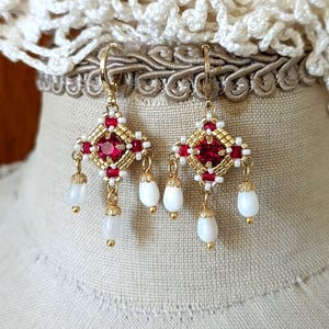 May include: Gold-toned earrings with a square, red gemstone center, surrounded by white and gold beads. Each earring has three dangling, white, teardrop-shaped beads. The earrings are displayed on a mannequin head.