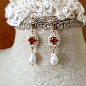 May include: A pair of gold-toned earrings with a red square center stone, surrounded by white beads and gold accents. Each earring has a dangling white teardrop-shaped pearl. The earrings are displayed on a mannequin.