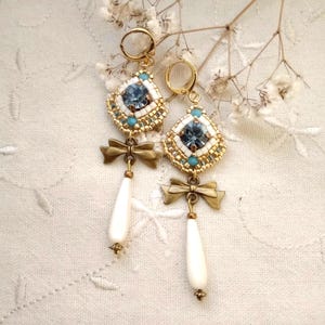 Peut inclure: Une paire de boucles d'oreilles dorées avec un cristal bleu, des perles blanches et une goutte de perle. Les boucles d'oreilles présentent un détail de nœud.