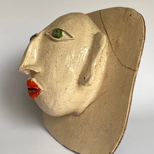 May include: A ceramic sculpture of a face with green eyes and red lips. The sculpture is white with a textured surface and has a crack on the side. The sculpture is mounted on a white ceramic base.