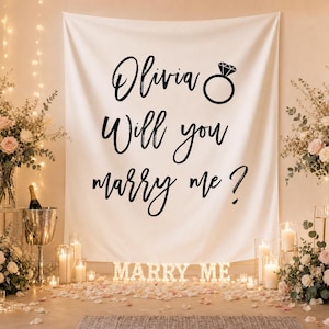 Op de afbeelding: Een witte stoffen achtergrond met de zwarte tekst "Olivia Will you marry me ?" en een icoon van een diamanten ring. Het doek wordt getoond in een kamer versierd met bloemstukken, kaarsen en verlichte "MARRY ME"-letters.