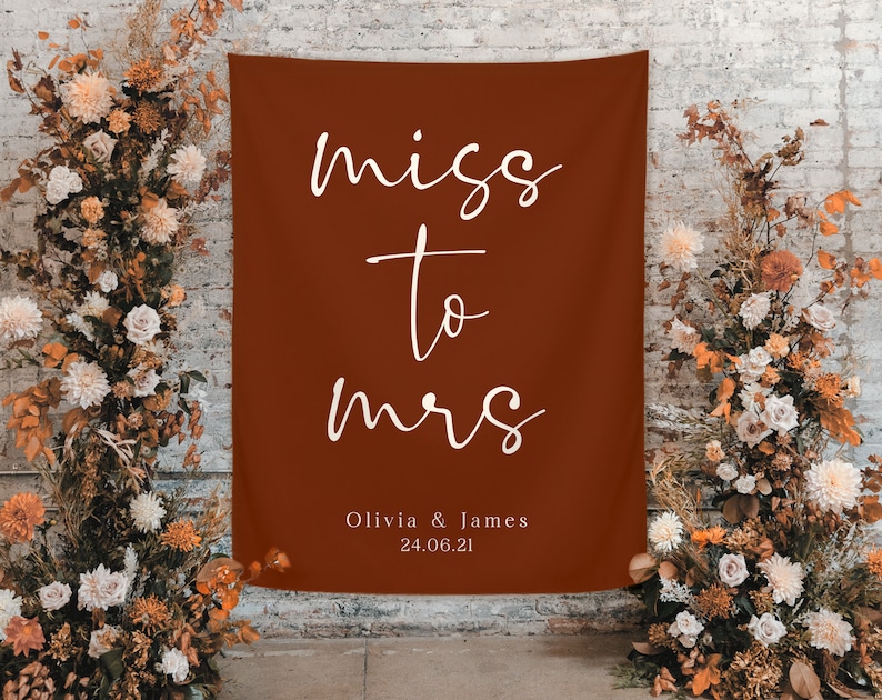 Pode incluir: Uma faixa de tecido cor de ferrugem com as palavras "miss to mrs" em escrita branca. Abaixo, os nomes "Olivia & James" e a data "24.06.21". A faixa &eacute; rodeada por arranjos florais.