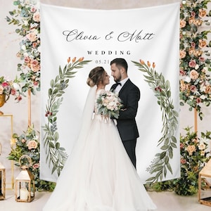 Peut inclure: Une toile de fond de mariage blanche avec les noms "Olivia & Matt" et la date "05.21" imprimés dessus. La toile de fond est décorée d'une couronne florale et de verdure. Un couple s'embrasse, la mariée tenant un bouquet.