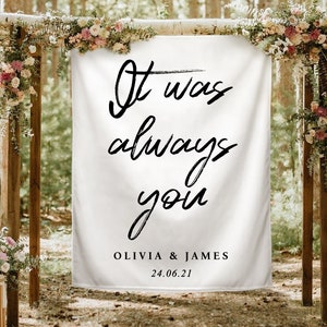Può includere: Uno striscione rettangolare in tessuto bianco con il testo in corsivo nero "It was always you", seguito dai nomi "OLIVIA & JAMES" e dalla data "24.06.21" in un carattere sans-serif, sospeso a una struttura in legno decorata con fiori.