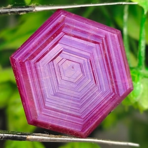 Peut inclure: Une pierre précieuse hexagonale, facettée, de couleur magenta et violet, est tenue par une pince à épiler devant un fond de feuillage vert. La gemme présente des motifs géométriques concentriques. Cette pierre est pour la joaillerie.
