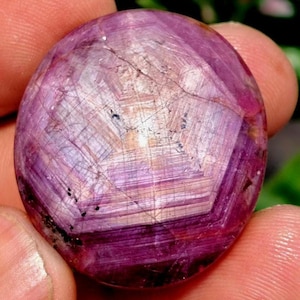 Puede incluir: Una gema de rubí pulida y redonda con un patrón en forma de estrella. La gema muestra un color rojo-púrpura intenso, con líneas internas que crean un efecto de estrella. La piedra se sostiene en una mano sobre un fondo verde borroso.