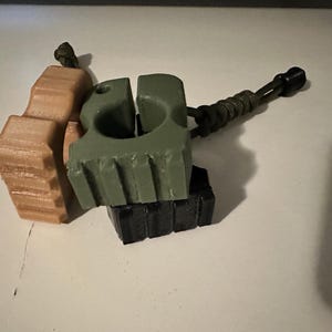 May include: Three small, geometric objects in shades of tan, olive green, and black. The green object has a hole and a cord attached. The objects have a textured, ridged design. The objects are likely 3D printed.