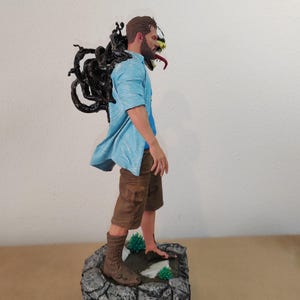以下が含まれることがあります： 黒と白の男性のアクションフィギュア。背中に黒いシンビオットの生き物がいます。フィギュアは青いシャツ、茶色のショートパンツ、茶色のブーツを履いています。フィギュアは緑の植物のある石の台座の上に立っています。