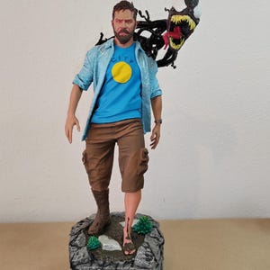 以下が含まれることがあります： ひげを生やした男性のフィギュアで、青いシャツに黄色の円、茶色のショートパンツを着用しています。口の大きな黒と赤の生き物が背後から現れています。フィギュアは、灰色の岩の台座の上に立っており、台座には「LEGEND」と書かれています。