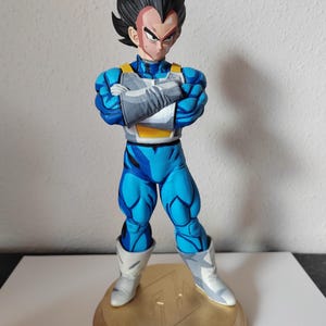 Puede incluir: Una figura de acción de Vegeta de Dragon Ball Z, de color azul y blanco. La figura está de pie con los brazos cruzados y tiene una expresión seria en su rostro. La figura está de pie sobre una base dorada con el logotipo de Dragon Ball Z.