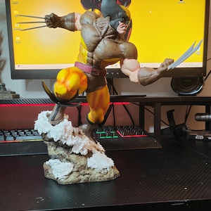 Peut inclure: Une figurine d'action de Wolverine en brun et jaune avec des griffes déployées, debout sur une base rocheuse. La figurine est posée dans une posture d'action dynamique, avec une jambe levée et l'autre fermement plantée sur la base. La figurine porte un costume brun et jaune avec une ceinture et un masque. La figurine est placée sur un fond noir.