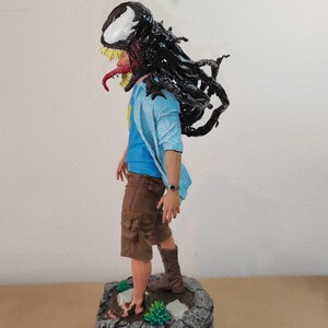 以下が含まれることがあります： 黒と白のキャラクターのフィギュア。頭と上半身を黒いシンビオートで覆っている。青いシャツと茶色のショートパンツを着用し、緑の植物のある岩の台座の上に立っている。