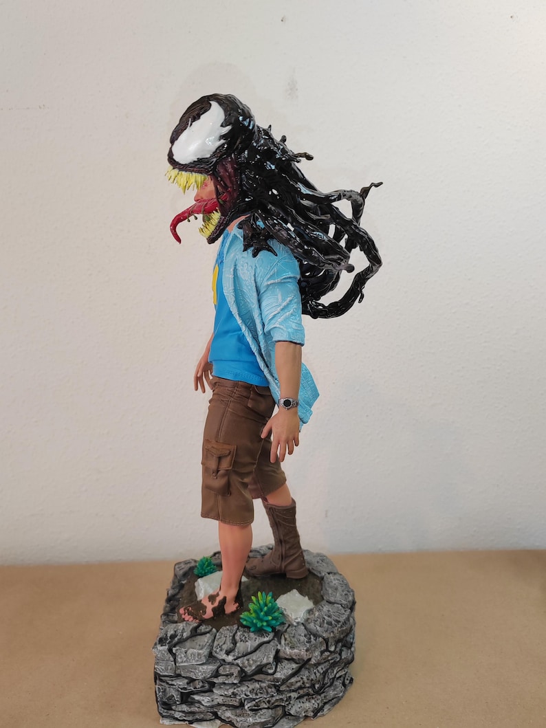 以下が含まれることがあります： 黒と白のキャラクターのフィギュア。頭と上半身を黒いシンビオートで覆っている。青いシャツと茶色のショートパンツを着用し、緑の植物のある岩の台座の上に立っている。