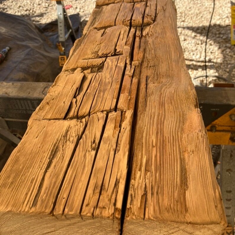 May include: Close-up of a weathered, light brown wooden beam with visible knots and grain patterns. The wood has a rustic and aged appearance.