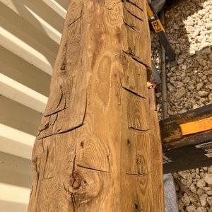 May include: A close-up of a large, weathered, wooden beam with a natural, rustic finish. The beam has a rough, textured surface with knots and grain patterns. The wood is a light brown color with darker brown streaks.