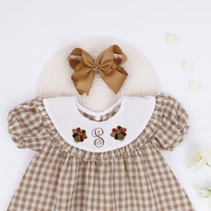 May include: A brown and white gingham dress with a white collar featuring embroidered turkey designs and the letter "G". A matching brown bow sits atop a straw hat. The dress has short, puffed sleeves.