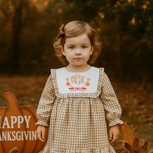 May include: A young child wearing a brown and white checkered dress with a white collar featuring an embroidered monogram and pumpkins. The dress has long sleeves and a ruffled hem. The child is wearing white socks and brown shoes, with a brown bow in their hair. Pumpkins and fall decor surround the child.