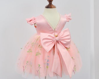 May include: A pink tulle dress with a satin bodice and a large pink bow. The dress has floral embroidery and a pearl trim around the neckline.