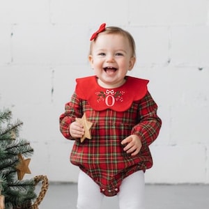 Può includere: Un bambino indossa un pagliaccetto a quadri rossi con un colletto rosso decorativo e un fiocco rosso abbinato. Il pagliaccetto ha maniche lunghe e leggings bianchi. Il bambino tiene in mano una stella di legno e sorride. A sinistra c'è un piccolo albero di Natale in un cesto intrecciato.