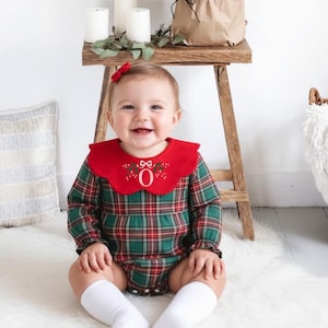 Può includere: Un bambino in un pagliaccetto a quadri rossi e verdi con un colletto rosso e il numero "0" ricamato. Il bambino è seduto su un tappeto bianco, sorridendo. Un piccolo albero di Natale è su uno sgabello di legno.