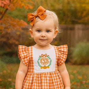May include: A toddler in an orange and white gingham dress with a pumpkin and floral design on the bodice. The outfit includes a matching bow headband, knee-high socks with bows, and brown Mary Jane shoes. The background features autumn foliage.