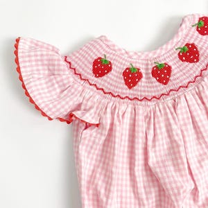 May include: A pink and white gingham romper with red trim and embroidered strawberries. The romper features a ruffled sleeve and a smocked neckline with three embroidered strawberries. The fabric is a light pink and white checkered pattern.