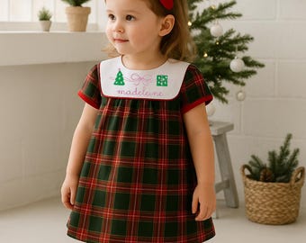 Könnte beinhalten: Ein junges Kind trägt ein grün-rotes kariertes Kleid mit einem weißen Kragen, der mit einem Weihnachtsbaum, einem Geschenk und dem Namen "Madeline" bestickt ist. Das Kind trägt rote Schuhe, weiße Socken mit roten Schleifen und eine rote Haarschleife.