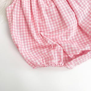 May include: A pink and white gingham romper with a red ruffled neckline and sleeve trim. The fabric is gathered at the neckline and the leg openings. The romper is made of a lightweight fabric.
