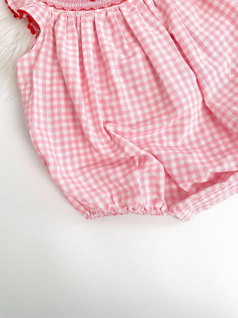 May include: A pink and white gingham romper with a red ruffled neckline and sleeve trim. The fabric is gathered at the neckline and the leg openings. The romper is made of a lightweight fabric.