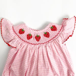 May include: A pink and white gingham baby dress with a ruffled neckline and sleeves. The neckline is embroidered with red strawberries. The dress has a red trim around the neckline and sleeves.