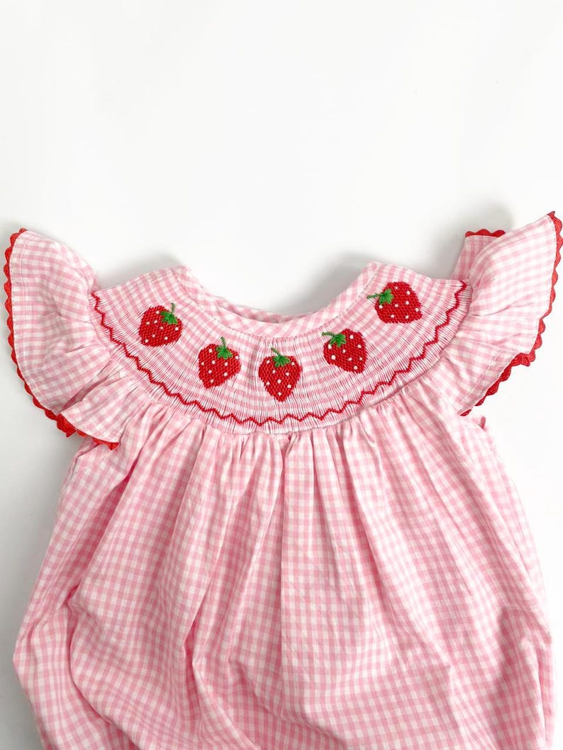 May include: A pink and white gingham baby dress with a ruffled neckline and sleeves. The neckline is embroidered with red strawberries. The dress has a red trim around the neckline and sleeves.
