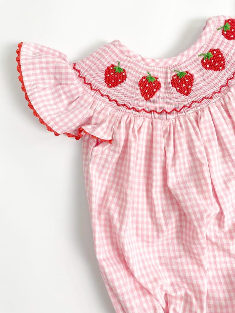 May include: A pink and white gingham romper with red trim and embroidered strawberries. The romper features a ruffled sleeve and a smocked neckline with three embroidered strawberries. The fabric is a light pink and white checkered pattern.